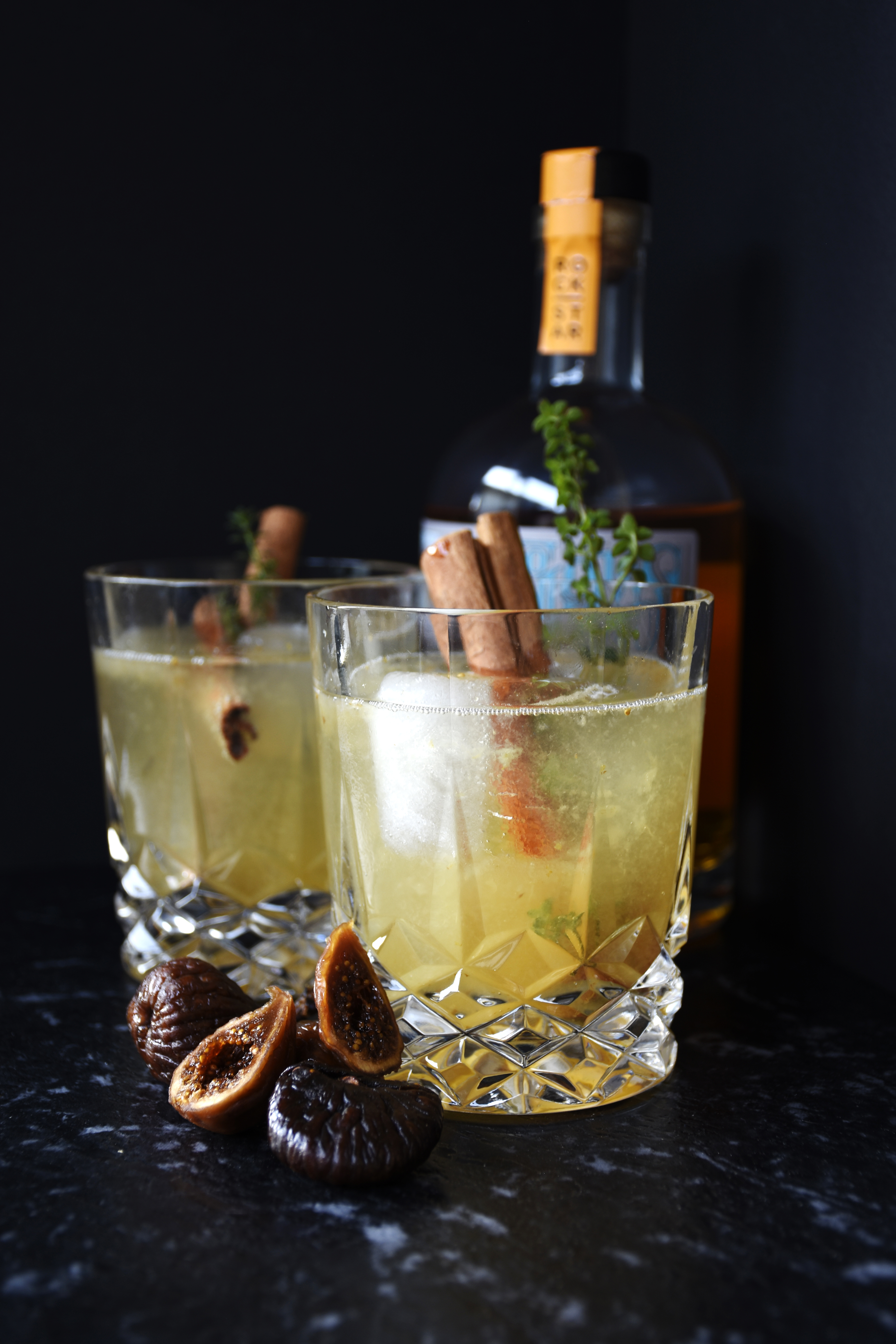 Two yellow coloured cocktails with cinnamon sticks and rosmary garnish showing out of the top in a short crystal glass against a black backdrop with dried figs sitting in front and a liquor bottle in the background